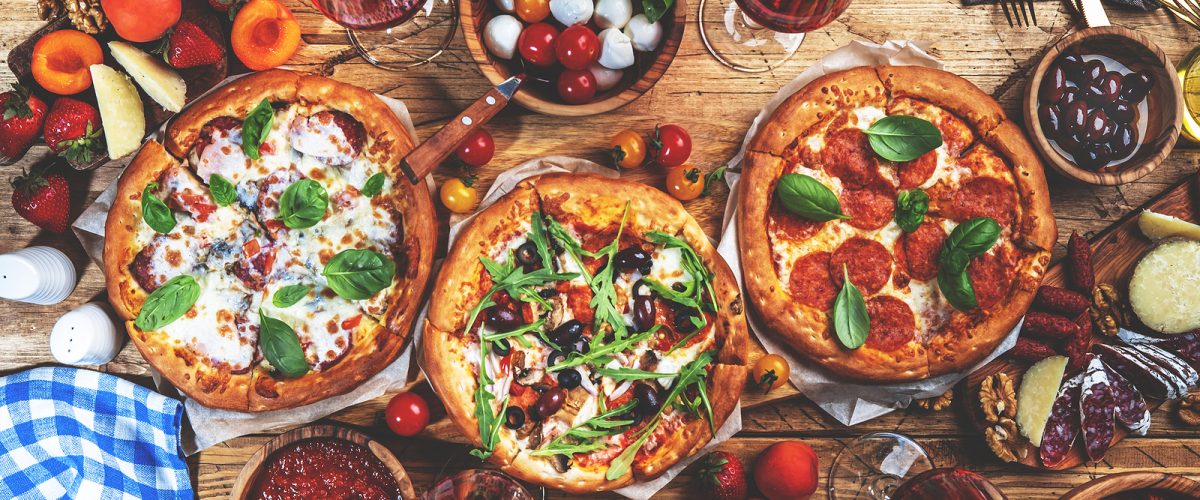 Pizza party table. Top view glasses with red wine, rustic wooden table with hot pizzas, italian appetizers, salads, cheese, fruits and berries. Family lunch with fast food
